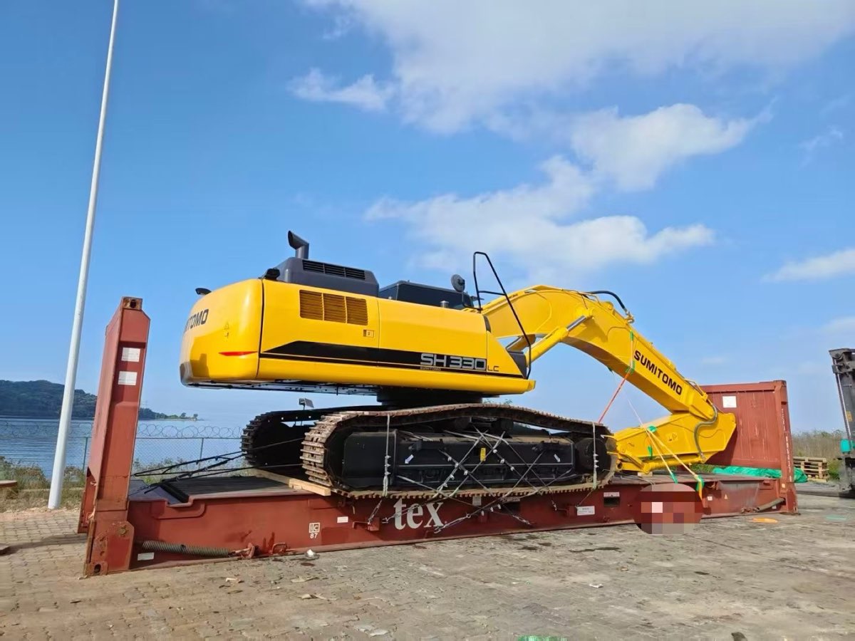 A yellow tracked excavator lashed to a red flat-rack platform at a port, ready for dispatch
